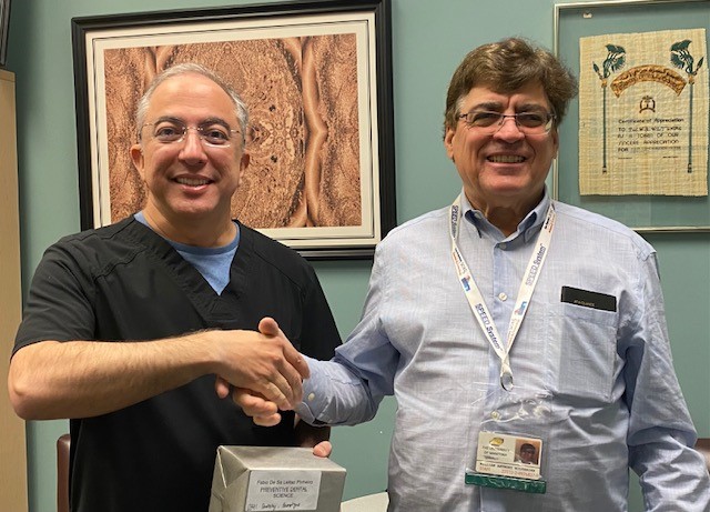 Dr. Wiltshire and Dr. Pinheiro shake hands. Dr. Pinheiro is holding a small box. 