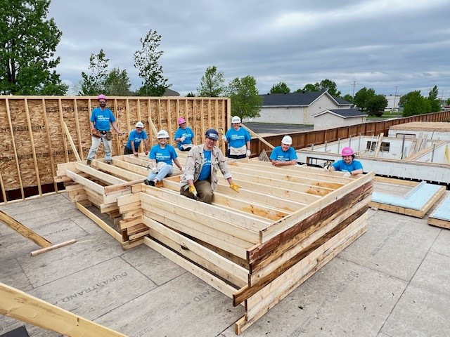 Eight people wearing hard hats sit and stand on wooden construction material. 