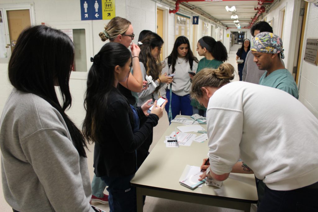 10 people stand around a table. One person has a swab in their mouth and another person is filling out a form. 