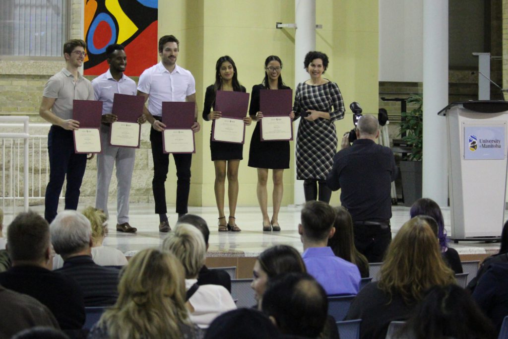 Five students hold certificates and pose with Dr. Anastasia Kelekis-Cholakis on a stage. A photographer is taking their photo. 