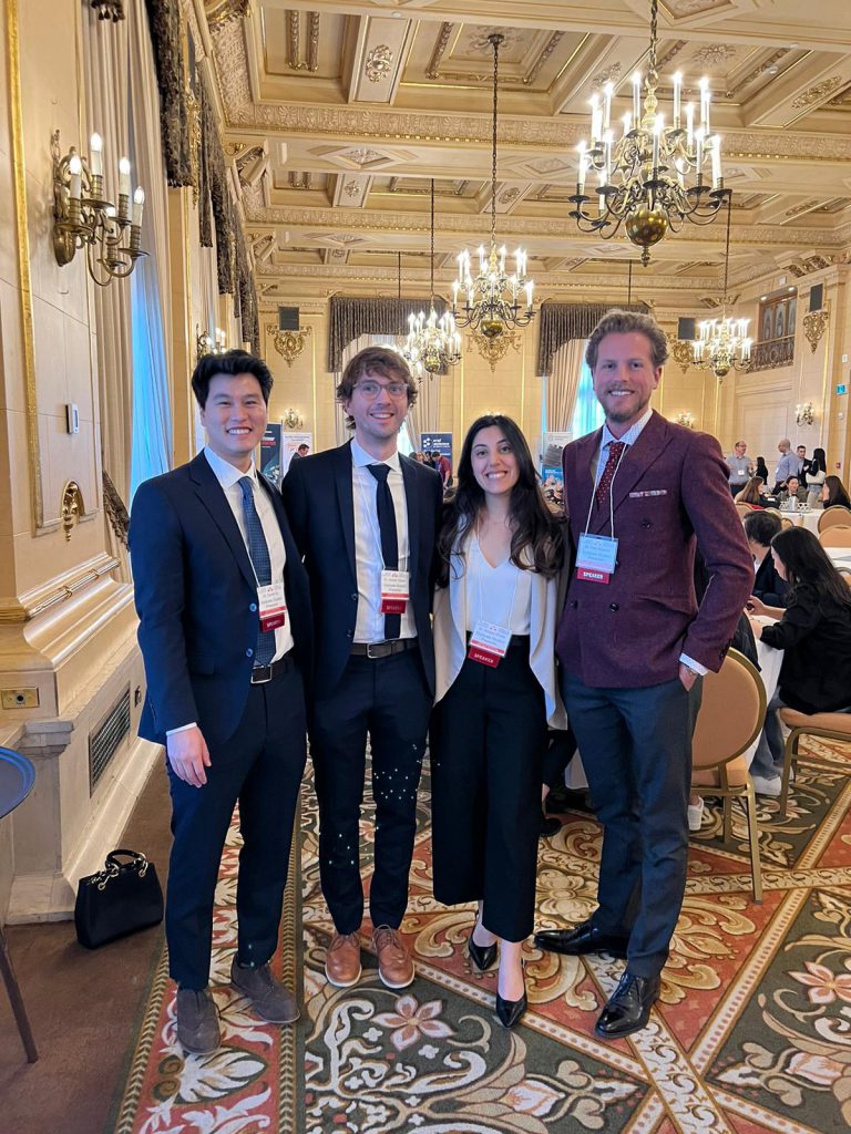 Four people pose in a grand ballroom featuring a fancy ceiling and chandeliers. 