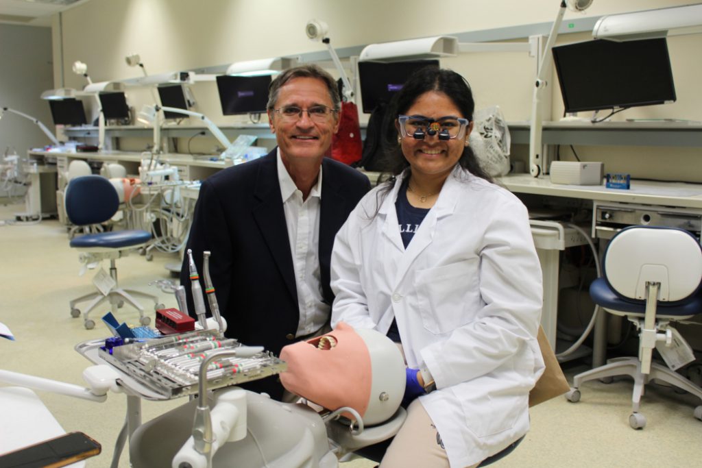 An alumnus poses with a dental student in a lab. A manikin is in front of them along with dental equipment. 