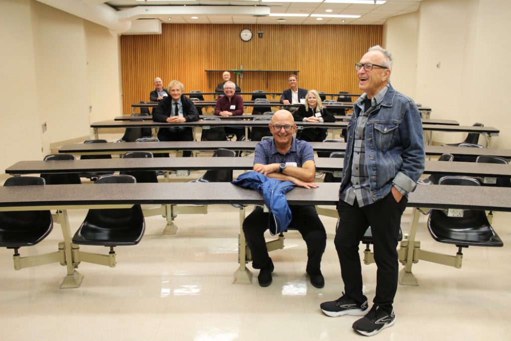 Seven people sit at desks. One person stands at the front of the classroom. 