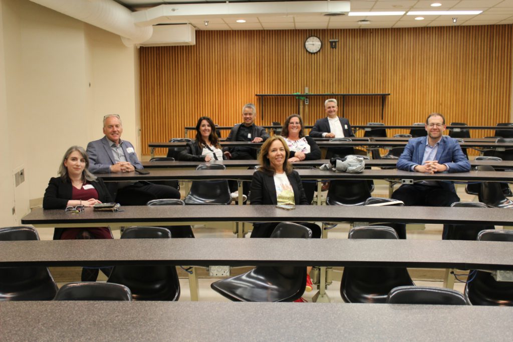 Eight people sit at desks in a classroom. 