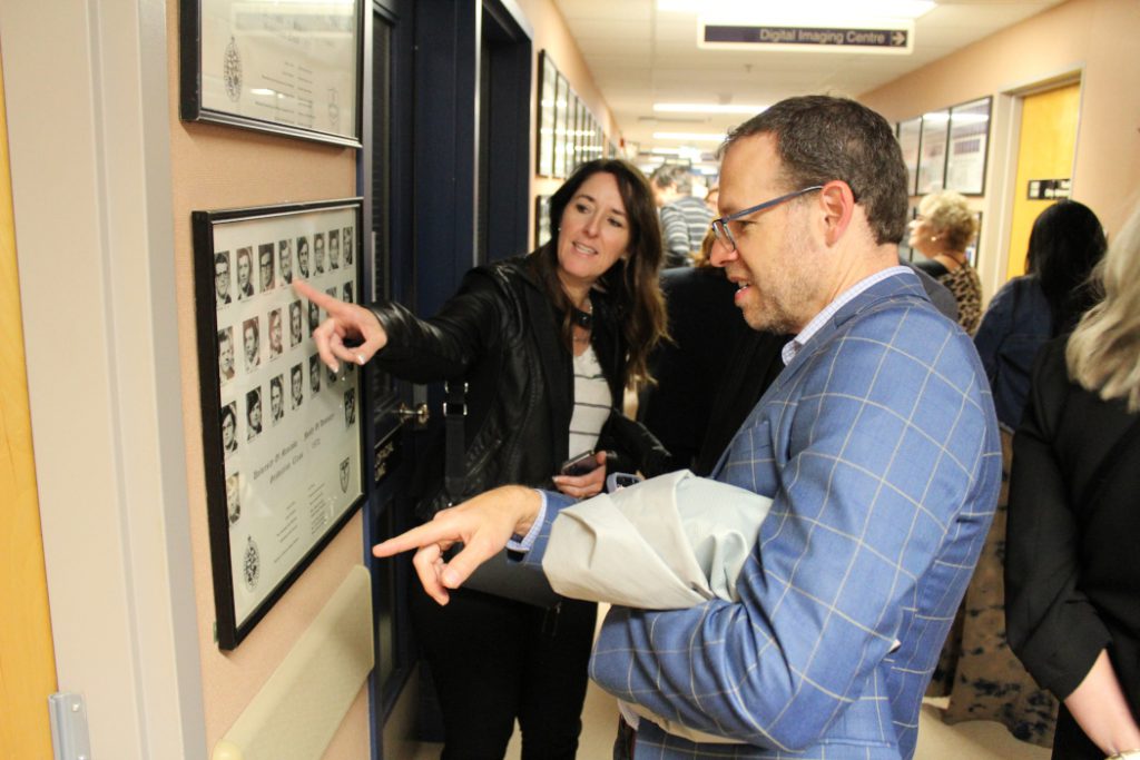 Two alumni point to graduation class photos on a wall. 