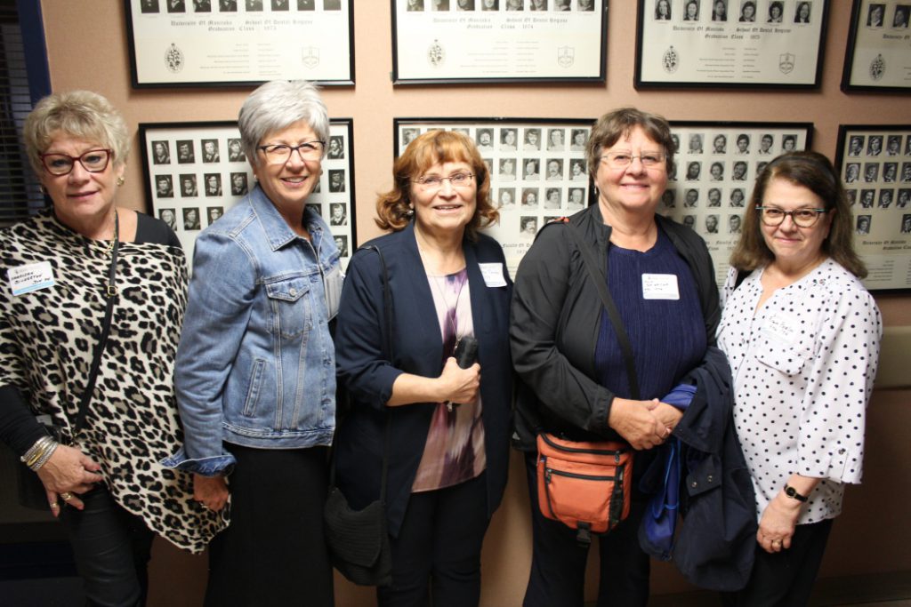 Five people pose for a photo in front of graduation class photos on a wall. 