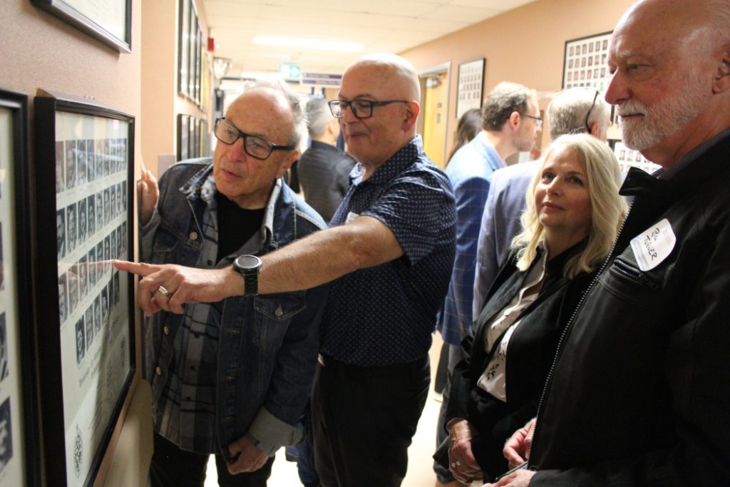 Four alumni look at graduation class photos on a wall. One alumnus points to a photo. 