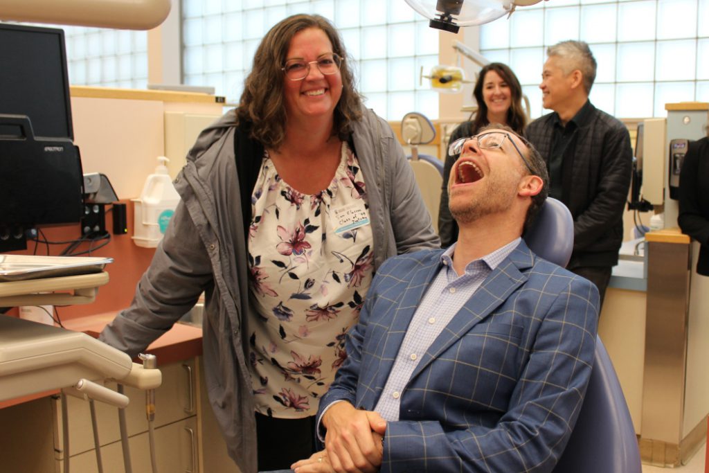 One alumnus sits in a dental chair and has their mouth open. Another alumnus stands next to the chair smiling. 