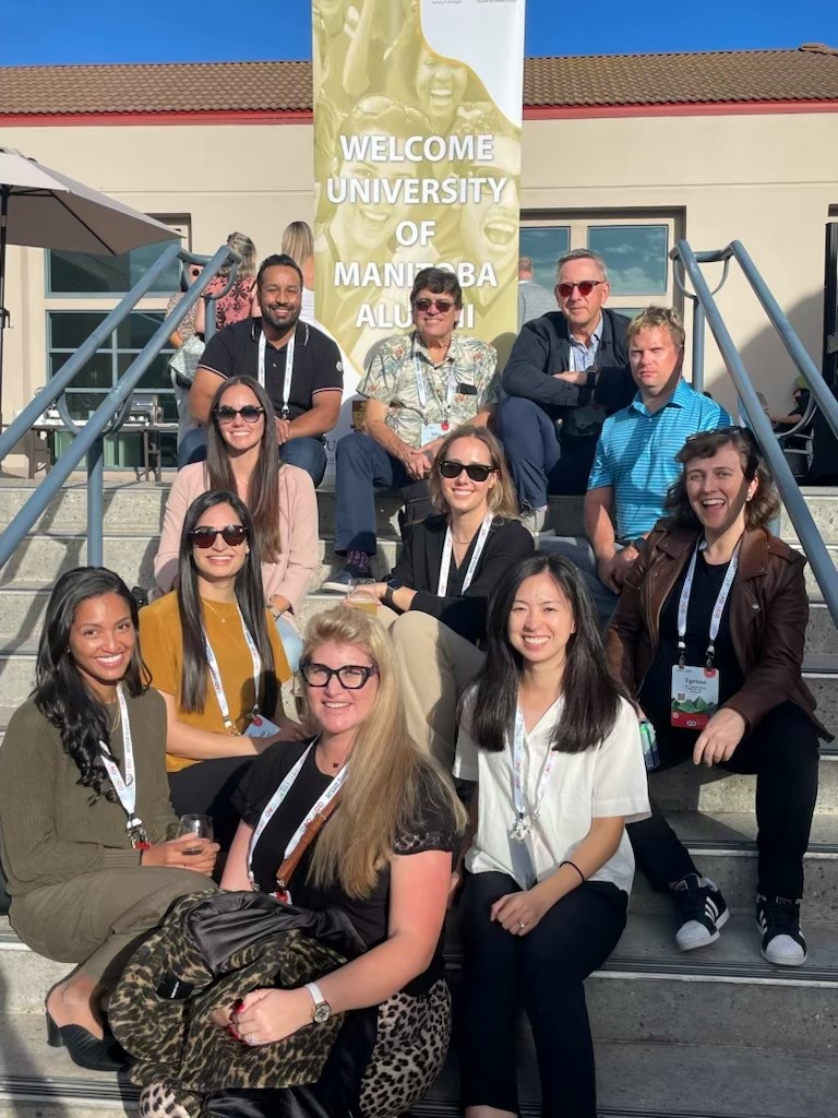 11 people pose for a photo sitting on steps. A banner at the top of the steps reads "Welcome University of Manitoba Alumni." 