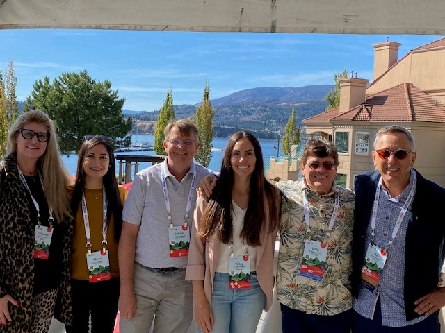 Six people pose for a photo. A lake, trees and hills are in the background. 