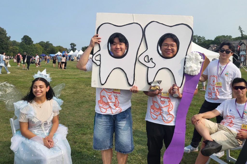 Two students hold a sign featuring two teeth. The teeth have holes in them that the students faces are seen through. One student is sitting down and is wearing a tooth fairy costume. 