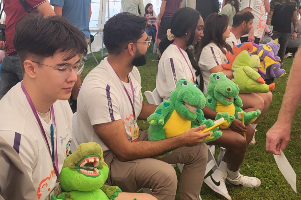 Five students are sitting in chairs. They are each holding a stuffed animal with teeth and a large toothbrush. 