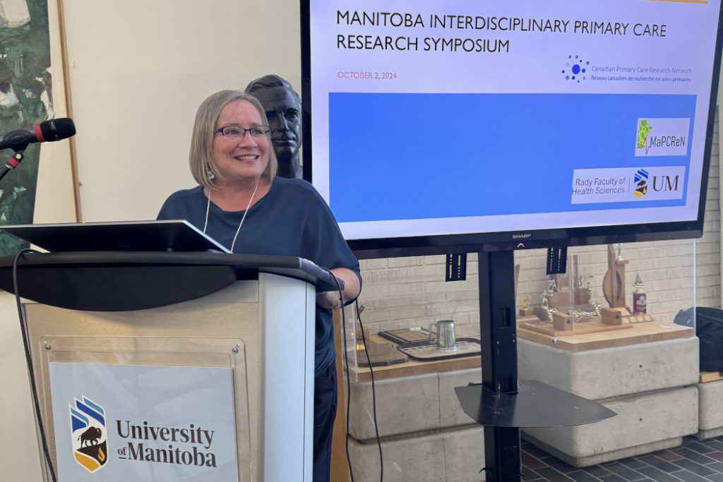 Dr. Gayle Halas stands at a lectern. Text on a screen behind her reads "Manitoba Interdisciplinary Primary Care Research Symposium." 