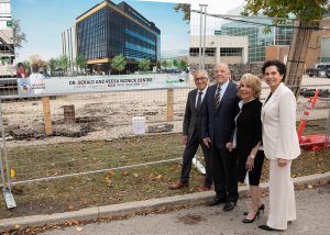 Four people stand next to a sign with a conceptual drawing of the new building. The sign is on a fence that surrounds the construction site. 