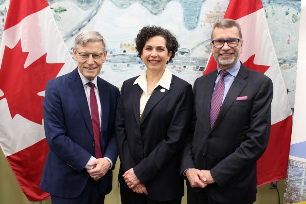 Three people stand in front of two Canadian flags. 