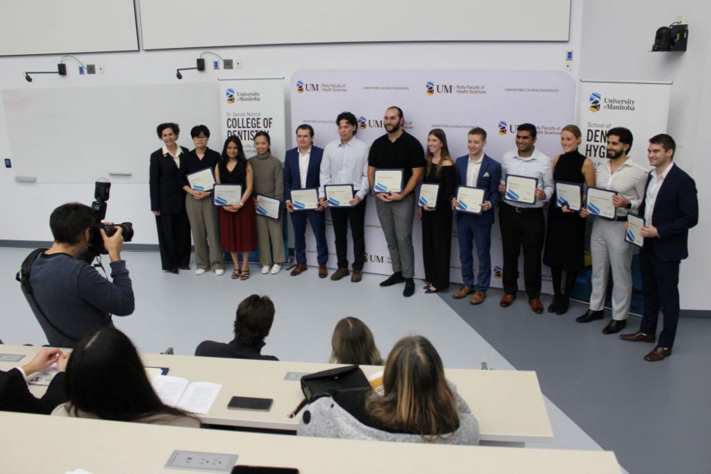 A photographer takes a photo of 12 students and Dr. Anastasia Kelekis-Cholakis. The students are holding certificates. 