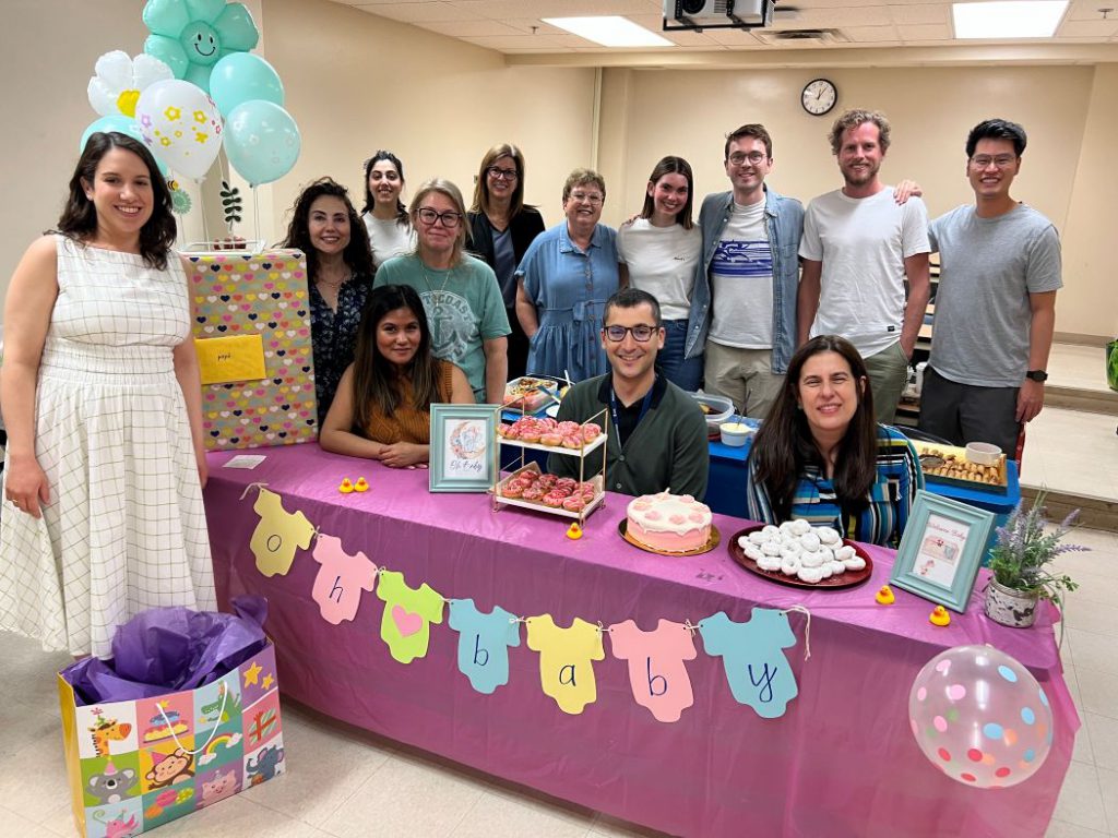 13 people pose for a photo. Gifts, balloons, and treats are on a table with a sign that reads "Oh baby". 