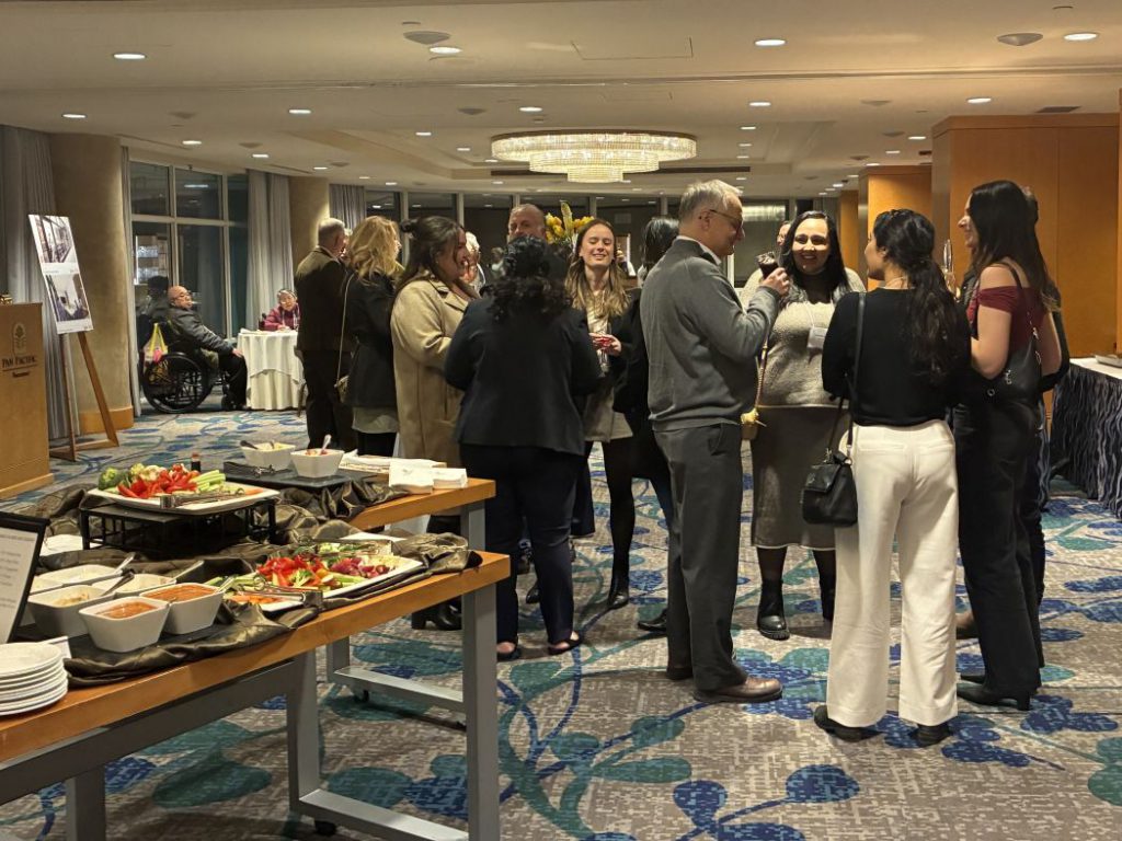 Groups of people talk and there is food on a table. 