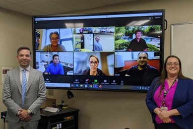 Two people stand next to a screen, which has six people on a Zoom call. 
