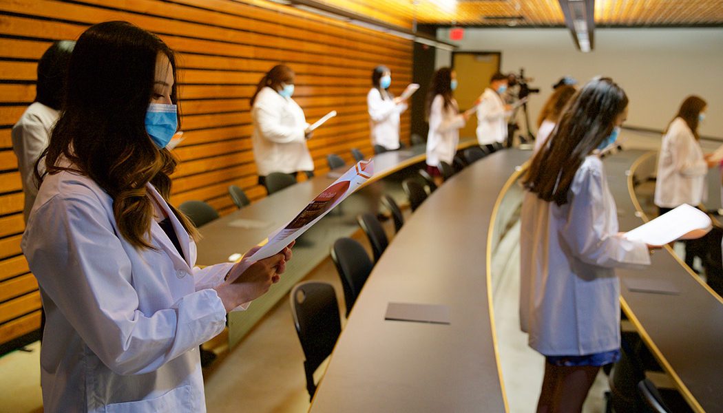 Pharmacy students wear white coats while holding papers and reading the Pledge of Professionalism.