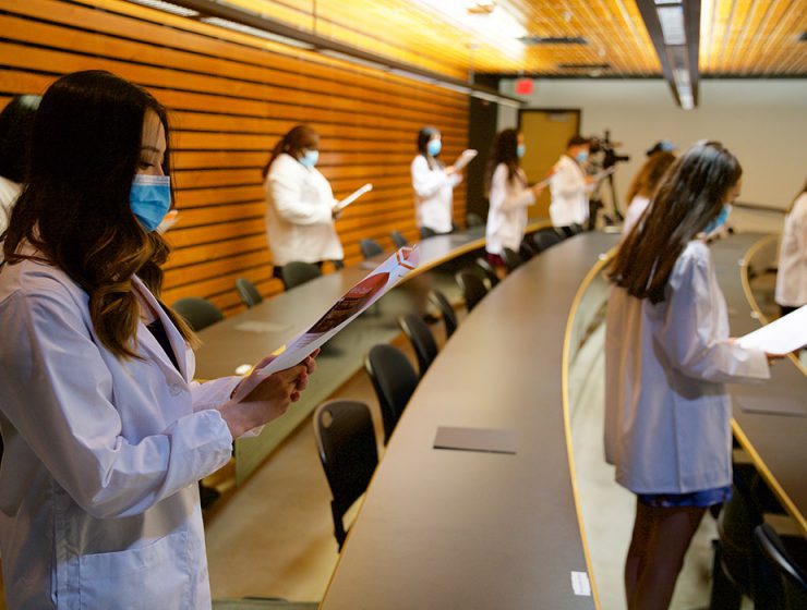 Pharmacy students wear white coats while holding papers and reading the Pledge of Professionalism.