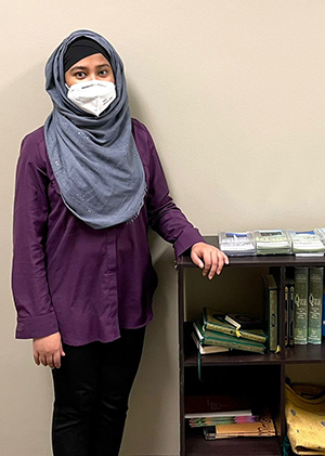 A Muslim student stands next to a bookshelf that contains copies of the Qur’an