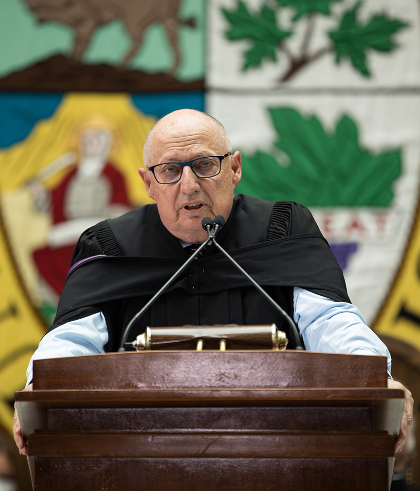 Dr. Brian Postl speaks at a lectern during a convocation ceremony.