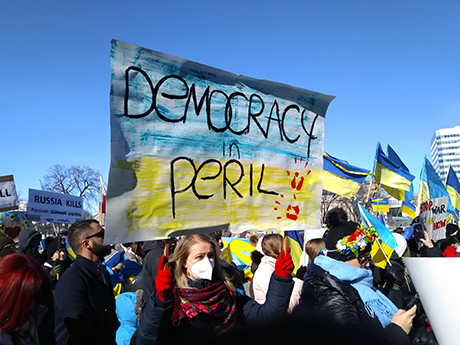 A group of protestors hold Ukrainian flags and signs. One reads "Democracy in Peril".