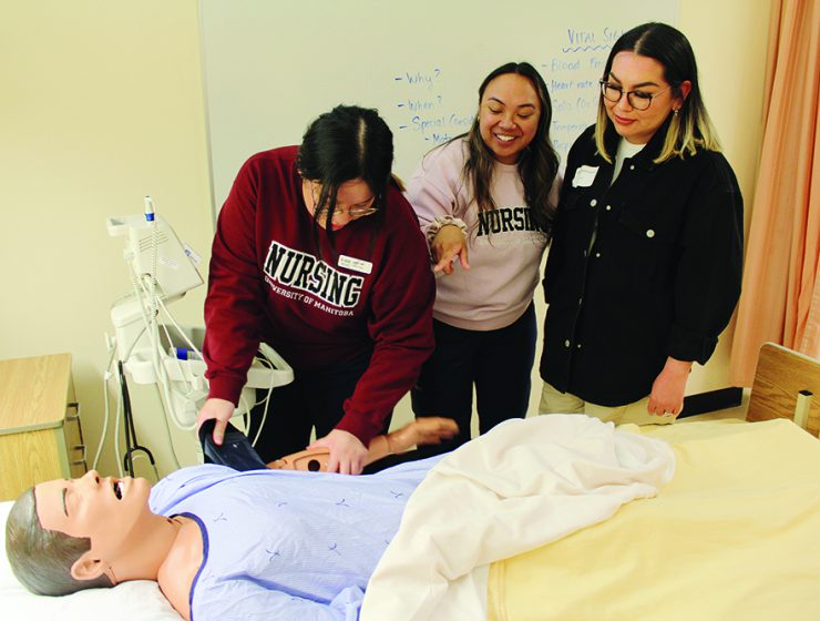 Two nursing students give a vital signs demonstration to an incoming student with a manikin in a hospital-style bed.