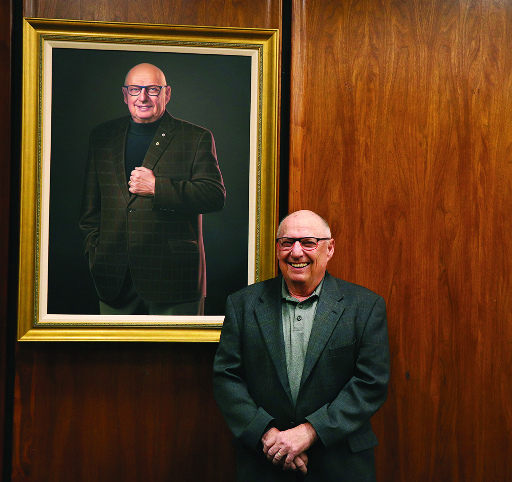 Brian Postl stands in front of his portrait, which hangs on a wood-paneled wall.