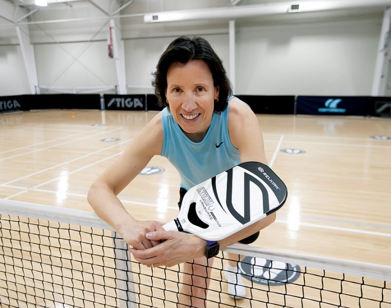 Sandra Webber holds a pickleball racquet and leans over a net.