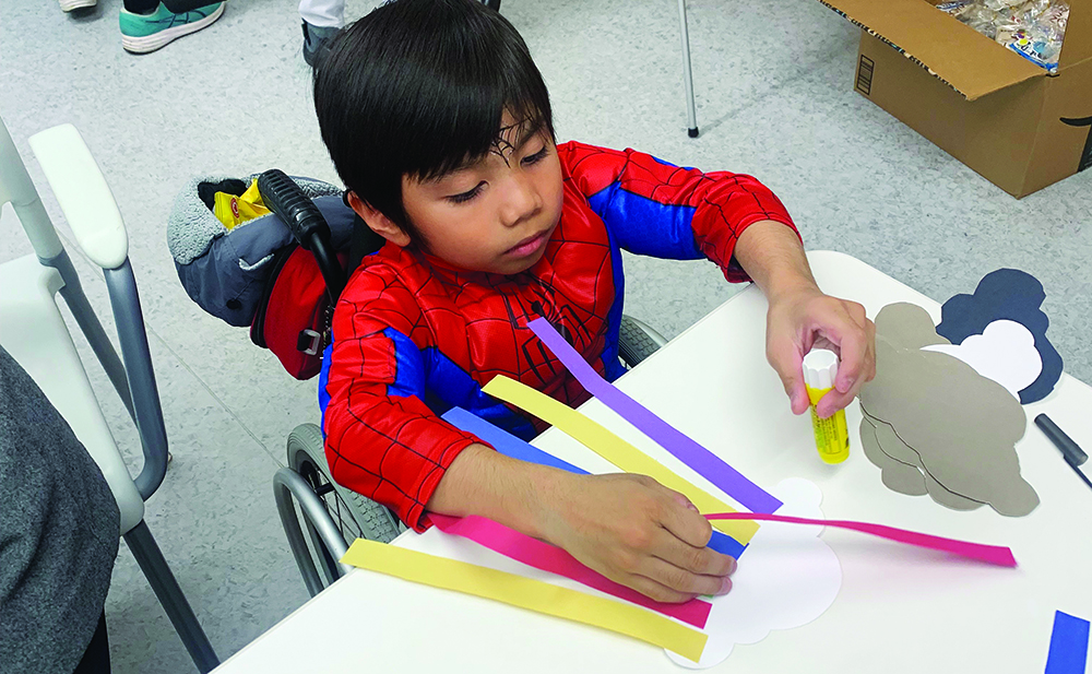 A young boy in a wheelchair, wearing a Spider-man costume, work at a craft table.