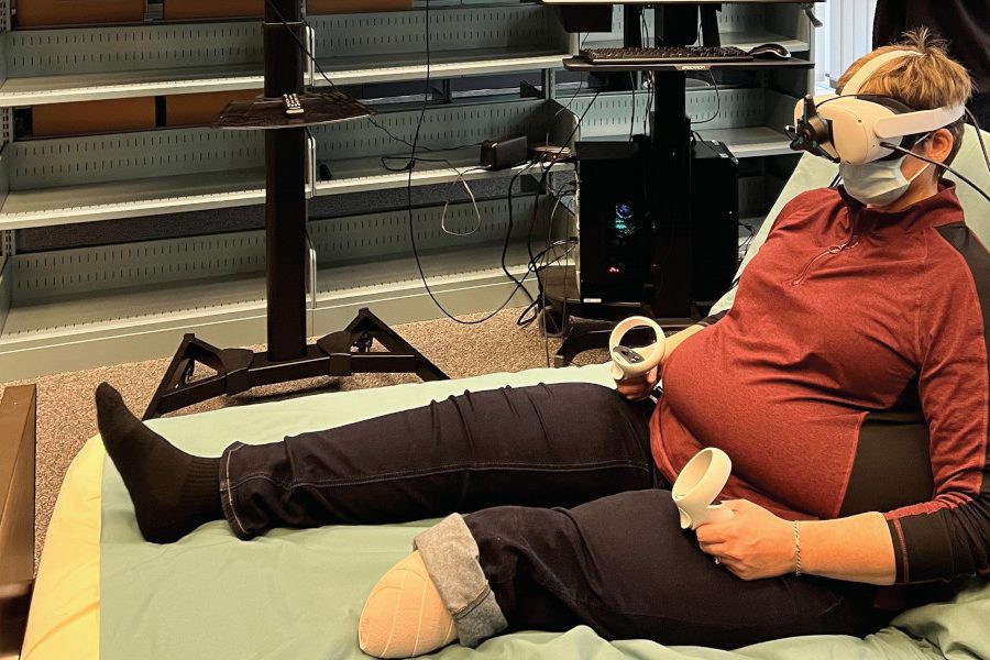 A patient partner sits up on a bed and is wearing a VR headset and is holding two VR controllers.