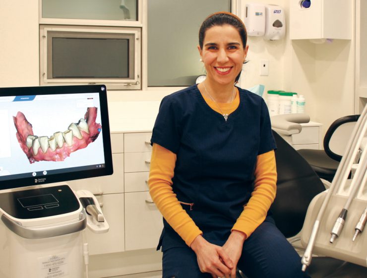 Dr. Hoda Hosseini sits on a dental chair. A 3D image of a lower set of teeth and gums is on a screen next to her.