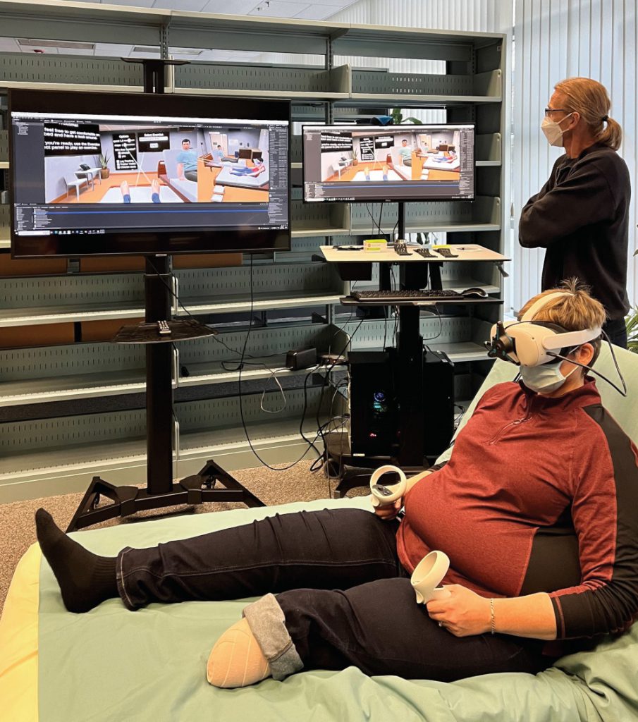 A patient partner sits up on a bed and is wearing a VR headset and is holding two VR controllers. The VR environment is on two screens to the side of the patient partner. 