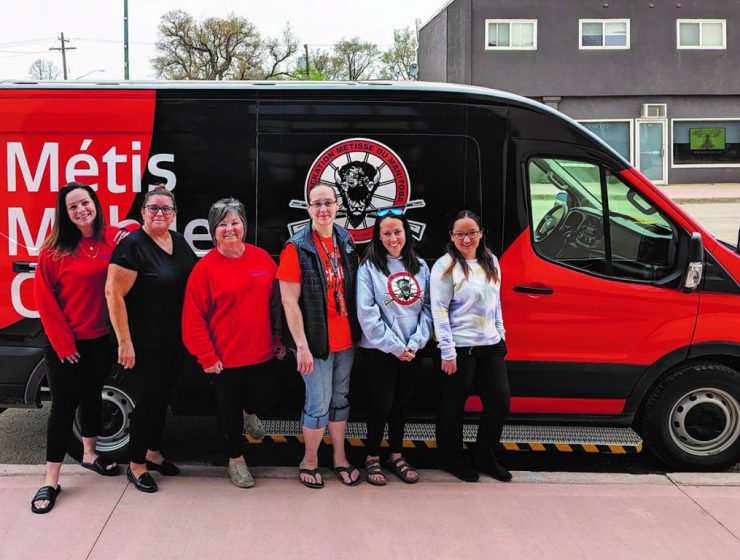 A group of six people stand in front of a red and black van branded with a Manitoba Metis Federation logo.