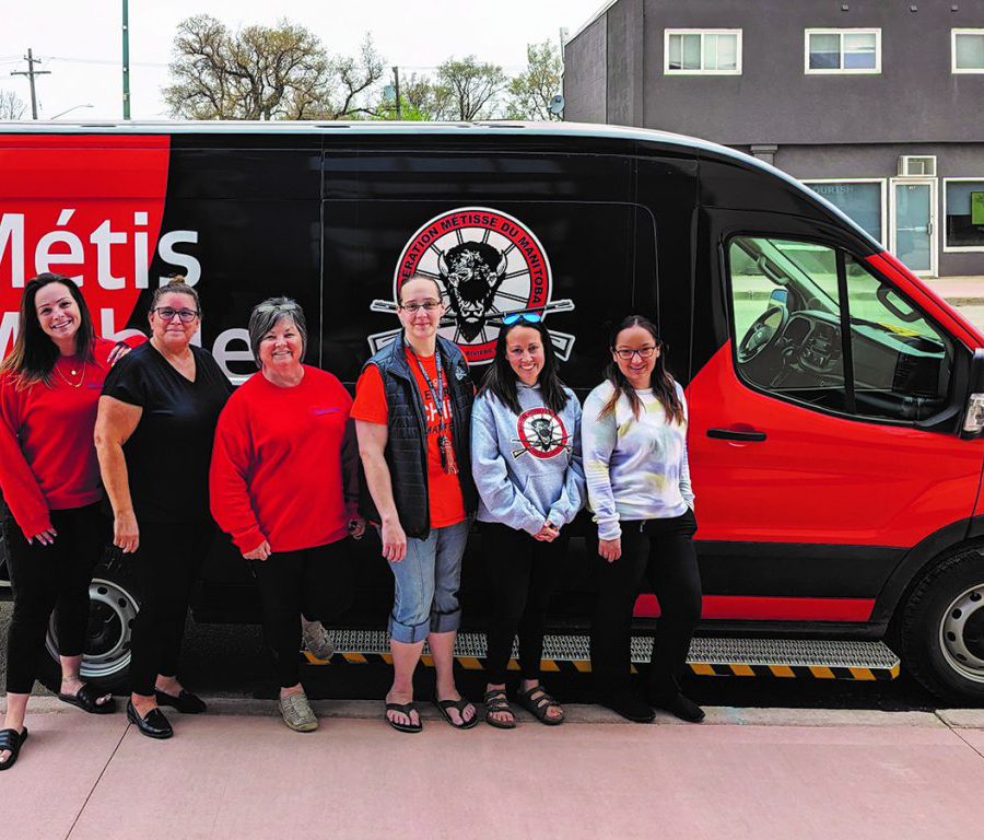 A group of six people stand in front of a red and black van branded with a Manitoba Metis Federation logo.