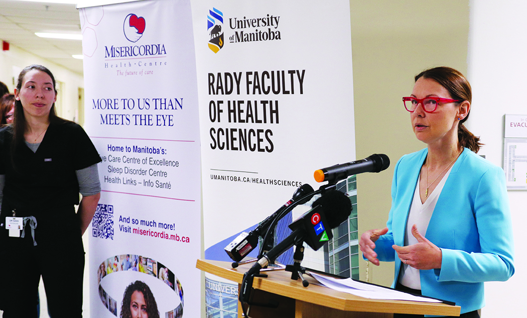 Nancy Porhownik stands in front of a podium in front of banners for the Rady Faculty of Health Sciences and Misericordia Health Centre. Sarah-Jane Sass stands to the side.