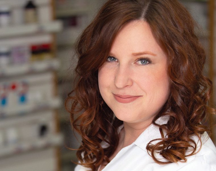 Portrait of Alia Marcinkow in a pharmacy. She is wearing a pharmacist's white coat.