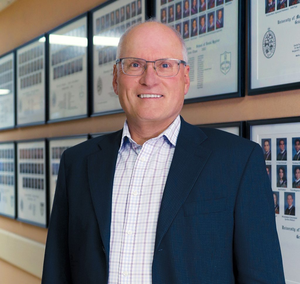 Portrait of Dr. John Perry. Dentistry student graduation photos are in frames on the wall behind him.