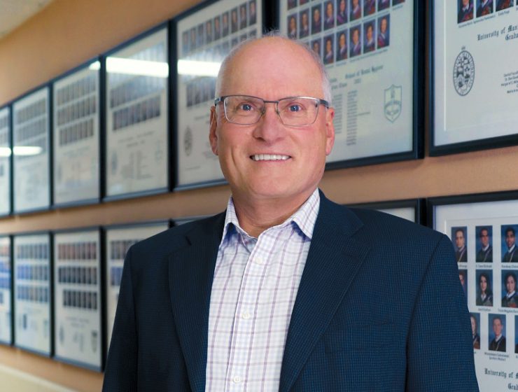 Portrait of Dr. John Perry. Dentistry student graduation photos are in frames on the wall behind him.