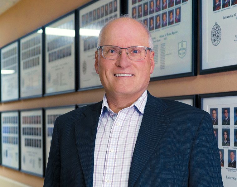 Portrait of Dr. John Perry. Dentistry student graduation photos are in frames on the wall behind him.