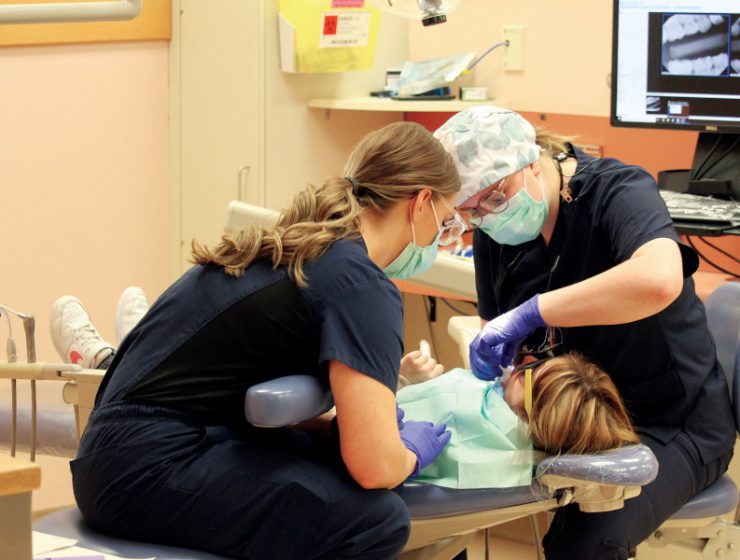 Two students perform a dental procedure on a child laying in a dental chair.