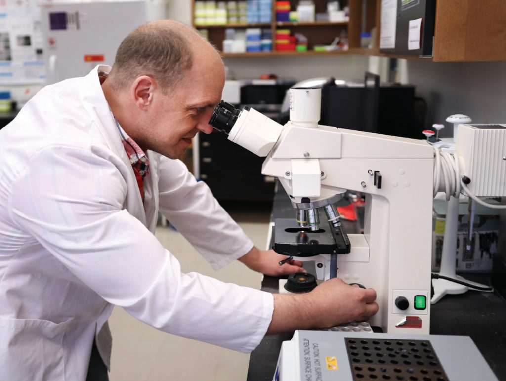 Dr. Jason Kindrachuk looks into a large microscope. He is wearing a lab coat. 