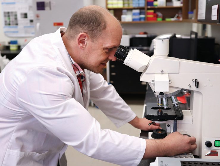 Dr. Jason Kindrachuk looks into a large microscope. He is wearing a lab coat.