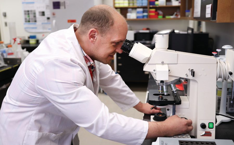 Dr. Jason Kindrachuk looks into a large microscope. He is wearing a lab coat.