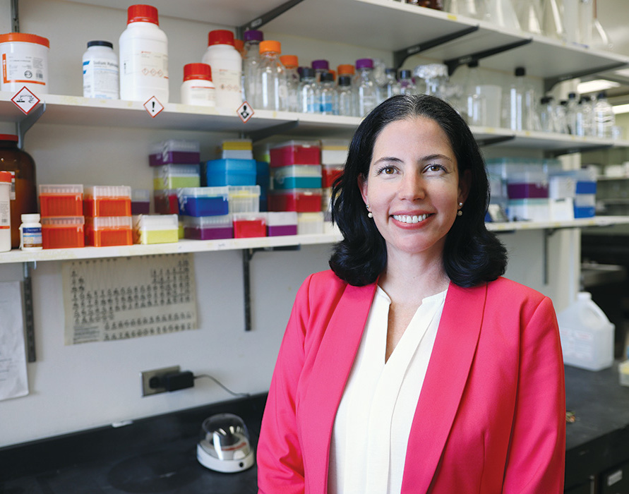 Portrait of Dr. Zulma Rueda in her lab. Containers used for scientific research sit on three shelves behind her. 