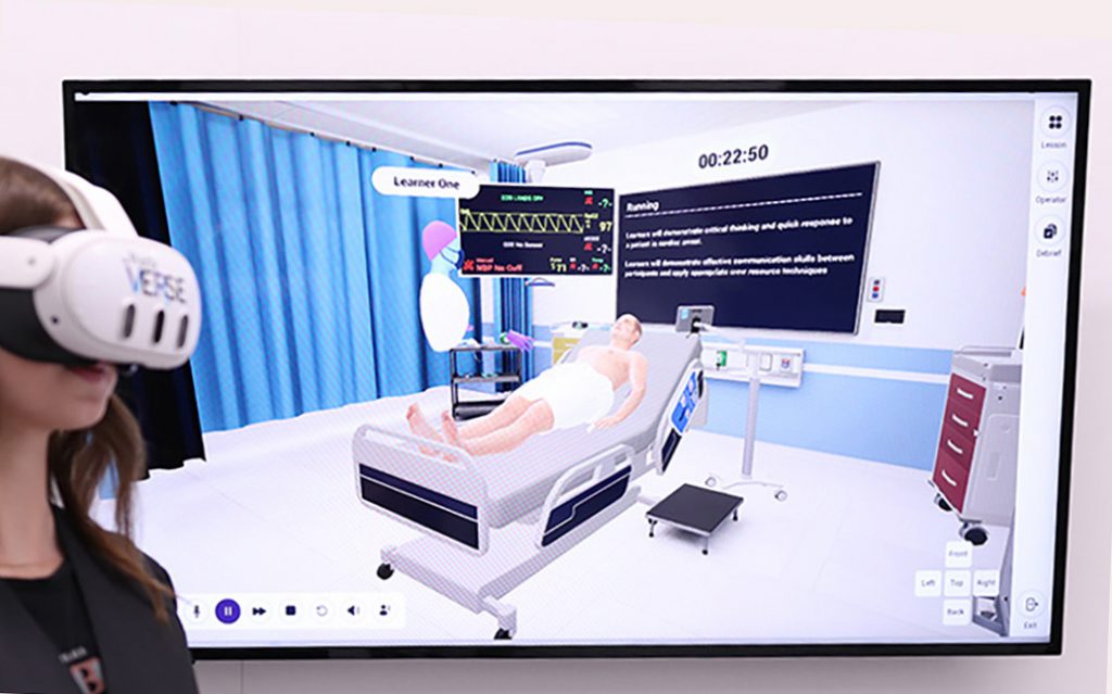 A person wearing a virtual reality headset stands in front of a TV showing a simulated hospital environment witha patient in a bed and an avatar labeled 'Leaner One.'