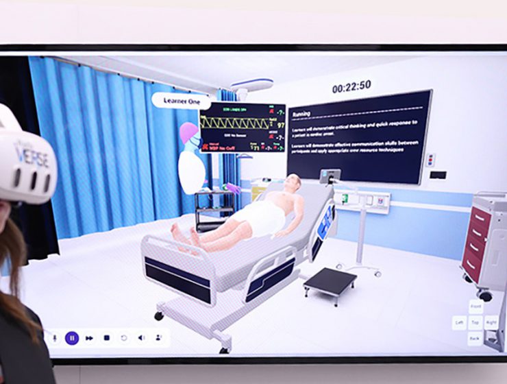 A person wearing a virtual reality headset stands in front of a TV showing a simulated hospital environment witha patient in a bed and an avatar labeled 'Leaner One.'