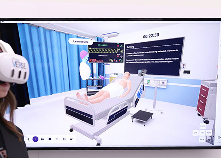 A person wearing a virtual reality headset stands in front of a TV showing a simulated hospital environment witha patient in a bed and an avatar labeled 'Leaner One.'